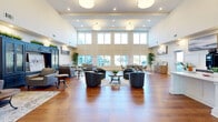 Located at 6039 Seminole Trail in Barboursville, Virginia, this single-floor community clubhouse offers a generously scaled, open-plan gathering space designed for both comfort and functionality. The defining architectural feature is the dramatic double-height ceiling, which floods the interior with natural light through a sweeping wall of oversized windows that frame views of the surrounding outdoor grounds and courtyard areas.

Rich hardwood flooring runs throughout the entire space, lending warmth and continuity across the various functional zones. The main lounge area is anchored by multiple seating arrangements — including plush leather sofas and contemporary swivel chairs — grouped around coffee tables atop layered area rugs, creating inviting conversation areas. Elegant wainscoting panels line the lower walls, adding a refined, classic character that complements the modern furnishings.

A well-appointed kitchenette serves the space with dark wood cabinetry, open floating shelves, white quartz countertops, a stainless steel refrigerator, and a central island. A dedicated dining area accommodates a full table and chairs, bathed in natural light from floor-to-ceiling windows overlooking the exterior. An ornate wrought-iron mezzanine railing above adds an architectural accent, while recessed and pendant lighting ensures the space is well-lit throughout.