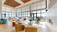 Located in Saint Petersburg, Florida, this generously sized rooftop amenity floor offers a sophisticated, open-plan layout designed for community gathering and elevated living. The main lounge area is anchored by soaring double-height ceilings and floor-to-ceiling windows that flood the interior with abundant natural light while framing unobstructed panoramic views of the surrounding cityscape, waterways, and lush greenery. A striking slatted wood ceiling feature adds warmth and architectural interest overhead, complementing the light hardwood flooring that runs throughout the space.

The interior is organized into distinct yet connected zones: a welcoming entry foyer with curated artwork and seating, a spacious main lounge with multiple conversation areas, a vibrant dining section accented by a bold tropical-inspired wall mural, and a well-appointed kitchen for hosting and catering. High-top seating and bar areas provide additional flexible options for gathering.

Multiple wraparound terraces extend the usable area outdoors, each featuring glass-paneled railings to preserve the elevated views. Outdoor lounge and dining furniture on the terraces make them ideal for al fresco relaxation, with sweeping sightlines stretching across the Saint Petersburg skyline, neighboring pools, and open skies. The overall ambiance is bright, coastal, and contemporary throughout.