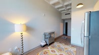 This single-floor studio unit offers a thoughtfully designed open layout that maximizes functionality within a compact footprint. The space immediately draws the eye upward with its distinctive coffered ceiling detail, adding architectural character and a sense of vertical spaciousness to the room.

The living and sleeping area occupies the main open space, anchored by warm wood-tone flooring and neutral gray walls. A large window at the far end floods the room with natural light and frames urban city views, creating a bright and airy atmosphere. A colorful area rug adds warmth and visual interest to the space.

The kitchenette is efficiently arranged along one wall, featuring white cabinetry that extends to the ceiling for ample storage, a stainless steel full-size refrigerator, a built-in microwave, an electric cooktop, a compact sink, and a dishwasher — all finished with stainless steel appliances for a clean, modern aesthetic.

The bathroom is neatly appointed with a wall-mounted white vanity with an integrated sink, a mirrored medicine cabinet, a shower with a curtain enclosure, and wood-tone flooring that complements the rest of the unit. Overall, the space maintains a clean, contemporary style suited for efficient urban living.