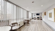 Located at 195 Clearview Ave in Ottawa, Ontario, this thoughtfully designed single-floor amenity space offers a welcoming and refined environment across a comfortable footprint. The space is divided into two distinct areas that flow seamlessly into one another, unified by light-toned luxury vinyl plank flooring and a cohesive neutral palette throughout.

The main lounge area is anchored by a classic white fireplace flanked by two upholstered armchairs and a coffee table, with a wall-mounted television above — creating a warm, residential focal point. Recessed lighting and textured grey wallcovering add depth and sophistication to the walls, which are further adorned with framed artwork and decorative accents in gold tones.

Floor-to-ceiling vertical blinds line an expansive window wall, flooding the space with abundant natural light and offering views of the surrounding tree-lined landscape. Beneath the windows, a row of bistro-style round tables with brass-toned legs and paired upholstered chairs creates an inviting setting for casual dining or conversation.

A fully equipped kitchen at one end of the space features stainless steel appliances — including a refrigerator and electric range — white shaker cabinetry, a subway tile backsplash, and clean white countertops, making it well-suited for group gatherings and catered events. The overall ambiance is modern, bright, and inviting.