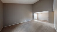 This single-story space is designed with a practical yet comfortable layout that accommodates a variety of living arrangements. The living area is a standout feature, characterized by dramatically vaulted ceilings that create an impressive sense of vertical space. Neutral gray walls and wall-to-wall carpeting in a warm beige tone establish a cohesive, understated aesthetic throughout. A sliding glass door with vertical blinds opens to an outdoor view, inviting natural light deep into the living space. An adjoining area, slightly separated by a change in ceiling height, features a ceiling fan and a window, offering a bright and functional secondary living zone.

The bedrooms are carpeted and finished in the same neutral palette, each equipped with windows that provide natural light and outdoor views. One bedroom includes a textured ceiling and a ceiling-mounted light fixture.

The kitchen is outfitted with dark cabinetry, speckled laminate countertops, a double stainless steel sink, and a dishwasher, with under-cabinet lighting and a window above the counter.

The bathroom includes a shower/tub combination with a decorative curtain and speckled flooring. A generously sized walk-in closet features built-in shelving and rod storage, offering ample organization capacity.