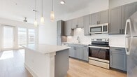 This well-designed one-bedroom, one-bathroom unit offers a thoughtfully organized single-floor layout. The open-concept kitchen and living area serves as the heart of the space, featuring light hardwood flooring throughout. The kitchen is outfitted with gray shaker-style cabinetry, a white tile backsplash, stainless steel appliances including a refrigerator, electric range, microwave, and dishwasher, and a central island with a clean white countertop. Pendant lighting above the island adds a modern touch. The living area benefits from natural light through glass doors leading to an outdoor area, with a ceiling fan overhead enhancing comfort. The bedroom is carpeted, with windows providing natural light and two closets offering ample storage. The elongated bathroom features gray cabinetry, a dual-sink vanity with white countertops, hardwood-style flooring, and access to an open shelving closet. Crisp white walls and recessed lighting throughout contribute to a clean, contemporary aesthetic.
