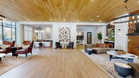 Located at 281 Cumberland Bend in Nashville, Tennessee, this single-floor community space offers a generously sized, open-plan layout designed with a refined yet welcoming aesthetic. The interior is immediately distinguished by its striking natural wood plank ceiling, which runs throughout the main area and adds warmth and visual continuity to the space.

The floor plan encompasses multiple distinct seating zones, including a lounge area anchored by a brick-surround fireplace with a mounted television, flanked by curved sofas and curated accent pieces. A second seating area near the entrance features rich cognac leather wingback chairs arranged around a side table, bathed in natural light from oversized black-framed windows that frame views of the surrounding greenery.

At the center of the space, a pair of mid-century style accent chairs flank a statement art piece displayed against a white brick accent wall, adding an artistic focal point. A dedicated kitchenette area provides practical functionality, showcasing a striking teal fish-scale mosaic tile backsplash, open wood shelving, white cabinetry, and stainless steel appliances.

Wood-look tile flooring flows seamlessly throughout, complementing the ceiling above. Large floor-to-ceiling glass entry doors and expansive windows flood the interior with natural light, enhancing the open, airy ambiance. The overall design balances contemporary style with warm, inviting materials.