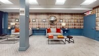 Located at 160 Temple St in New Haven, Connecticut, this thoughtfully designed single-floor space blends a library-inspired aesthetic with modern functionality. The interior is immediately defined by its striking floor-to-ceiling bookshelf wallcovering, which wraps around the room and creates a warm, intellectual ambiance throughout. A bold teal wainscoting runs along the lower portion of the walls, adding a refined architectural detail that contrasts elegantly with the patterned carpeting underfoot.

The space is organized into distinct functional zones. A central collaborative area features a large wooden communal table with industrial-style metal chairs, ideal for group work or meetings. Along one wall, a dedicated workstation zone includes desktop computers set atop a built-in teal desk, providing a productive environment for focused tasks. A pair of industrial metal shelving units flank this area, offering additional storage and display space.

The lounge area provides a comfortable retreat, furnished with gray sofas accented by vibrant orange throw pillows. Decorative round mirrors mounted above low teal credenzas, alongside table lamps and framed artwork, contribute to the space's curated, editorial character. Recessed panel ceiling lighting ensures the entire space is evenly and efficiently illuminated. The overall atmosphere is inviting, professional, and creatively stimulating.