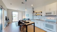 This contemporary single-floor residence showcases a clean, modern aesthetic throughout, with crisp white walls, light hardwood flooring, and an open-concept layout that maximizes the sense of space. The kitchen is thoughtfully designed with white cabinetry, a subway tile backsplash, gray quartz-style countertops, a full suite of appliances, and open wood shelving with black metal accents for added character. A built-in island with bar seating provides a casual dining option and serves as a natural transition into the living area. The living room is bright and welcoming, anchored by a large floor-to-ceiling sliding glass door that floods the space with natural light and offers open urban views. The bedroom continues the modern theme with plush carpet flooring and oversized windows on two walls, framing neighborhood views and allowing abundant daylight. The full bathroom features white cabinetry, a gray countertop vanity, and a combination tub and shower. A dedicated utility closet houses a stacked full-size washer and dryer alongside the HVAC system, providing practical in-unit convenience. The overall ambiance is bright, clean, and contemporary, reflecting a well-appointed urban living space.