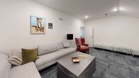 This single-floor space presents a well-appointed lounge area designed for comfort and casual gathering. The room is defined by clean white walls and textured blue-gray carpeted flooring, lending a calm, professional ambiance throughout.

The seating arrangement centers around a generously sized light gray L-shaped sectional sofa, accented with olive green and neutral throw pillows. A distinctive red leather wingback chair serves as a bold focal point, adding warmth and character to the otherwise neutral palette. Two mint green upholstered ottomans offer additional flexible seating near the room's entrance. A low-profile gray upholstered coffee table anchors the seating area, topped with books and decorative objects.

Lighting is provided by recessed ceiling fixtures supplemented by two floor lamps — a brass arc lamp with a black shade and a sleek black globe floor lamp — creating a layered, welcoming glow. An air purifier tucked into one corner reflects attention to comfort and functionality.

The walls are adorned with playful, dog-themed artwork, including a vibrant painted portrait of a Boxer dog and several smaller framed pieces, alongside a classic eye chart print that adds a quirky, memorable touch. A doorway leads to an adjacent room visible in the background, suggesting additional space beyond. The overall atmosphere is relaxed yet thoughtfully curated.