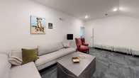 This single-floor space presents a well-appointed lounge area designed for comfort and casual gathering. The room is defined by clean white walls and textured blue-gray carpeted flooring, lending a calm, professional ambiance throughout.

The seating arrangement centers around a generously sized light gray L-shaped sectional sofa, accented with olive green and neutral throw pillows. A distinctive red leather wingback chair serves as a bold focal point, adding warmth and character to the otherwise neutral palette. Two mint green upholstered ottomans offer additional flexible seating near the room's entrance. A low-profile gray upholstered coffee table anchors the seating area, topped with books and decorative objects.

Lighting is provided by recessed ceiling fixtures supplemented by two floor lamps — a brass arc lamp with a black shade and a sleek black globe floor lamp — creating a layered, welcoming glow. An air purifier tucked into one corner reflects attention to comfort and functionality.

The walls are adorned with playful, dog-themed artwork, including a vibrant painted portrait of a Boxer dog and several smaller framed pieces, alongside a classic eye chart print that adds a quirky, memorable touch. A doorway leads to an adjacent room visible in the background, suggesting additional space beyond. The overall atmosphere is relaxed yet thoughtfully curated.