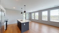 This single-floor residence presents a thoughtfully designed open-concept layout that seamlessly connects the kitchen and living area. The kitchen serves as the centerpiece, anchored by a substantial island with a white quartz countertop, an integrated sink, and striking slate-blue cabinetry. A pair of matte black pendant lights hang above the island, adding a modern industrial touch. The kitchen wall features white shaker-style upper cabinets, dark lower cabinets, a classic white subway tile backsplash, and stainless steel appliances including a range, over-the-range microwave, and a French door refrigerator. Warm-toned hardwood-style flooring extends throughout the open living space, which is bathed in natural light from a generous row of windows fitted with horizontal blinds. The bedroom continues the same flooring and neutral gray palette, offering corner windows on two walls and a ceiling fan with light fixture for added comfort. The full bathroom is finished with large dark-format floor tiles, a white subway tile tub surround, a white shaker vanity, and sleek matte black hardware and fixtures. A second bathroom mirrors this aesthetic with coordinating tile floors and a white vanity, completing the cohesive, contemporary design of the space.