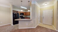 This single-floor residence is designed with a functional, open layout that connects its main living spaces seamlessly. The entryway, finished with tile flooring and crown molding, leads directly into the living and dining areas, which feature warm-toned carpet and crown molding throughout, contributing to a cohesive and polished aesthetic.

The kitchen is well-appointed with rich medium-toned wood cabinetry, dark countertops, and a full suite of black appliances, including a refrigerator, range, microwave, and dishwasher. A decorative diamond-pattern wine rack adds a distinctive detail above the upper cabinets. The kitchen opens to the living and dining areas via a raised breakfast bar with pendant lighting overhead, creating an inviting, connected atmosphere.

The bedroom is a generously sized, carpeted room equipped with a ceiling fan and a window that allows natural light to filter in. An attached bathroom features a cherry-toned vanity, tile flooring, and a large mirror. Adjacent to the bathroom, a dedicated laundry area with a washer and dryer provides added convenience. The overall color palette of warm yellows and neutral tones lends the space a comfortable, welcoming character throughout.