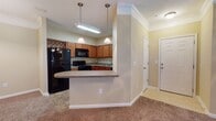 This single-floor residence is designed with a functional, open layout that connects its main living spaces seamlessly. The entryway, finished with tile flooring and crown molding, leads directly into the living and dining areas, which feature warm-toned carpet and crown molding throughout, contributing to a cohesive and polished aesthetic.

The kitchen is well-appointed with rich medium-toned wood cabinetry, dark countertops, and a full suite of black appliances, including a refrigerator, range, microwave, and dishwasher. A decorative diamond-pattern wine rack adds a distinctive detail above the upper cabinets. The kitchen opens to the living and dining areas via a raised breakfast bar with pendant lighting overhead, creating an inviting, connected atmosphere.

The bedroom is a generously sized, carpeted room equipped with a ceiling fan and a window that allows natural light to filter in. An attached bathroom features a cherry-toned vanity, tile flooring, and a large mirror. Adjacent to the bathroom, a dedicated laundry area with a washer and dryer provides added convenience. The overall color palette of warm yellows and neutral tones lends the space a comfortable, welcoming character throughout.