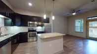 Located at 2223 Waterloo City Ln in Austin, TX, this single-story unit showcases a thoughtfully designed open-concept layout with modern finishes throughout. The kitchen serves as the centerpiece of the living space, featuring dark espresso cabinetry that extends to the ceiling, a white subway tile backsplash, and a full suite of stainless steel appliances including a range, microwave, and refrigerator. A freestanding island with a light granite countertop anchors the space and is illuminated by two pendant lights, creating an ideal spot for casual dining or meal prep. Wood-look flooring runs throughout the main living and kitchen areas, contributing to a warm, cohesive aesthetic. The open layout flows seamlessly from the kitchen into the living area, which features a ceiling fan and access to natural light from a nearby window. The bedroom is finished with plush carpeting, a ceiling fan, and a window that allows natural light to fill the room. The bathroom is clean and functional, offering a full tub-shower combination with white tile surround, a dark-vanity with light countertop, marble-look tile flooring, and a large mirror. Overall, the space presents a contemporary and well-appointed interior suitable for comfortable everyday living.
