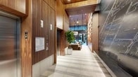 This elegant building lobby is a study in modern design, blending warm materials with contemporary architecture to create a welcoming and functional amenity floor. The entry corridor immediately impresses with its rich vertical wood slat paneling, terrazzo tile flooring, and a dramatic dark feature wall etched with an intricate city map graphic, lending an artistic and urban character to the space.

The corridor opens into a generously proportioned lounge area anchored by a full-wall built-in shelving unit crafted in warm wood tones, complete with integrated accent lighting and a large flat-screen display. Plush, earth-toned seating arrangements — including a modular sectional sofa, accent chairs, and organic-shaped coffee tables — create an inviting gathering space beneath soaring ceilings and a statement drum pendant light. Lush potted plants throughout add a natural, refreshing element to the interior.

Adjacent to the lounge, a well-equipped fitness center features modern cardio machines, including a treadmill and elliptical, with floor-to-ceiling glass doors that open directly to a private outdoor courtyard. The courtyard offers an intimate al fresco setting with a dining table, garden seating, lush greenery, and overhead string lights that lend a warm, convivial ambiance. Elevators with sleek finishes provide convenient vertical access throughout the building.