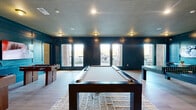 Located in Vestavia Hills, Alabama, this Game Center is a thoughtfully designed single-floor entertainment venue that offers a dynamic and inviting atmosphere for recreation and socializing. The space is defined by its bold deep teal paneled walls, which are accented by detailed wainscoting that runs throughout, lending the room a polished, upscale character. Light hardwood flooring contrasts beautifully against the rich wall color, while a distinctive textured ceiling with warm-toned flush-mount fixtures adds visual interest overhead.

The main area is generously proportioned, accommodating a full-size pool table at its center, a foosball table, and a dedicated shuffleboard table along the perimeter. Multiple large flat-screen televisions are mounted on the walls, making the space ideal for watching sports or entertainment while gaming. A built-in bar-height counter lined with leather-upholstered stools provides a comfortable viewing and socializing station. Bistro-style seating arrangements offer additional spots for guests to gather.

Natural light pours in through oversized windows and glass panel doors along one wall, offering views of the surrounding brick exterior and outdoor areas. The overall ambiance is energetic yet refined, striking a balance between a lively game room and a sophisticated lounge environment.