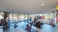 This single-floor fitness center is designed to provide a comprehensive workout experience in a bright, open environment. Large, floor-to-ceiling windows wrap around a significant portion of the space, flooding the interior with natural light and offering unobstructed views of the surrounding greenery and landscaping. The light-toned flooring and white walls contribute to a clean, airy aesthetic throughout.

The cardio zone is well-appointed with treadmills, elliptical trainers, and stationary spin bikes, several of which are equipped with personal screens for an interactive workout experience. Rubber exercise mats are positioned beneath the equipment to provide stability and floor protection. A dedicated strength training area features a multi-station cable machine and an adjustable weight bench, catering to a full range of resistance exercises.

A functional training section houses a variety of medicine balls in assorted weights, stability balls, and resistance accessories, all neatly organized on a rack. A wall-mounted flat-screen television is positioned for easy viewing from multiple workout stations. Recessed lighting in the textured ceiling supplements the abundant natural light. Sanitation stations and a waste bin are conveniently placed, reflecting a commitment to cleanliness and user comfort throughout the facility.