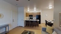 This single-floor residence showcases a thoughtfully designed open-concept layout that integrates the kitchen, dining, and living areas into one cohesive space. The kitchen is a standout feature, outfitted with warm wood-toned cabinetry that extends to the upper walls, providing generous storage. Black appliances, including a refrigerator and microwave, offer a sleek contrast against the light-colored countertops. A peninsula with a built-in sink serves as the kitchen's centerpiece, complemented by stylish black metal bar stools that invite casual dining and conversation. Flush-mount ceiling lights illuminate the kitchen area warmly. Light wood-toned flooring runs throughout, lending continuity and a clean, contemporary aesthetic to the entire space. The living area, partially visible, features a relaxed seating arrangement anchored by a glass-top coffee table. A dedicated dining area with dark wood chairs is positioned to one side, adding a defined sense of purpose within the open floor plan. White walls and neutral tones throughout create a bright, airy atmosphere, while a ceiling fan adds a practical touch to the overall design.