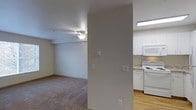This single-floor residential unit offers a clean, functional layout designed for comfortable everyday living. The living area is finished with wall-to-wall carpeting and features a ceiling fan with integrated lighting, lending a practical and cozy ambiance to the space. Neutral-toned walls throughout create a bright, cohesive feel that easily accommodates a variety of furnishing styles. A structural column partially divides the living area from the kitchen, maintaining an open sense of flow between the two spaces. The kitchen is equipped with white appliances, including a freestanding range, over-the-range microwave, and refrigerator, all complemented by white cabinetry and light-toned countertops. The kitchen flooring transitions to hardwood-style material, providing a clean contrast to the carpeted living space. Recessed fluorescent lighting illuminates the kitchen area efficiently. Multiple wall switches are conveniently positioned throughout, and the overall layout reflects a straightforward, well-maintained design. The space is well-suited for those seeking a tidy, move-in ready environment with a simple and practical aesthetic.