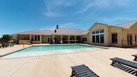 Located at 501 S Loop 250 Frontage Rd in Midland, Texas, this single-story amenity complex offers a well-appointed outdoor recreation area designed for both relaxation and social gatherings. The centerpiece of the space is a generously sized swimming pool, framed by a broad concrete sun deck lined with dark wood lounge chairs, perfect for taking in the West Texas sunshine under open blue skies. The pool area is enclosed by a wrought iron fence, with a safety ring station visible poolside.

The main building features a classic Texas Hill Country aesthetic, constructed with natural stone masonry and topped with a standing-seam metal roof. Large multi-pane windows allow ample natural light into the interior spaces. A covered outdoor patio extends from the building, supported by painted columns and equipped with ceiling fans for year-round comfort. This shaded gathering space includes dining tables and chairs, wall-mounted lantern lighting, and a stainless steel built-in gas grill, making it ideal for outdoor entertaining.

The interior areas visible in the space appear to be in a transitional or renovation state, with exposed walls and minimal furnishings. Surrounding multi-story residential buildings with balconies are visible in the background, providing context for the community-oriented nature of this amenity space.
