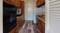 Located at 8802 Three Chopt Rd in Henrico, Virginia, this single-floor unit offers a practical and functional layout throughout its living spaces. The galley-style kitchen is efficiently designed with warm wood cabinetry lining both sides, providing ample storage. Dark wood-look flooring runs the length of the kitchen, complementing the black appliances, including a gas cooktop and wall oven, alongside a white refrigerator and stainless steel sink with a window-facing orientation near the entry door.

The main living area is generously proportioned, featuring soft neutral carpeting and a series of large windows along two walls that invite abundant natural light and offer pleasant views of mature surrounding trees. A pendant light fixture anchors the space, and a louvered closet door adds a practical storage element. The open, airy feel of this room makes it a versatile and welcoming central space.

The bathroom retains a classic mid-century character, showcased by its distinctive light blue subway tile work extending along the walls, a full soaking tub with shower, a vanity with sink, and wood-look flooring that ties back to the kitchen's aesthetic. Overall, the unit presents a cohesive, functional living environment.