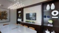 This single-floor space showcases a thoughtfully designed interior with a strong emphasis on contemporary style and refined finishes. The focal point of the living and dining area is a dramatic feature wall clad in light-toned stone or tile paneling, framed by rich dark wood cabinetry. A wall-mounted flat-screen television is seamlessly integrated into this feature wall, flanked on either side by built-in shelving and display cabinetry with glass-fronted panels, offering both functional storage and an opportunity to showcase decorative accents. Below the television, a low credenza provides additional surface space, currently styled with greenery for a touch of organic contrast against the dark wood tones. The dining area sits in close proximity, anchored by a smooth, light-colored table surrounded by contemporary seating. Overhead, a cascading chandelier composed of individual pendant bulbs serves as a sculptural lighting element, adding visual drama and warmth to the space. The overall palette — combining crisp whites, warm wood tones, and neutral stone — creates a cohesive, upscale ambiance throughout.