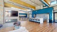 Located at 955 W Clairemont Ave in Eau Claire, Wisconsin, this expansive single-floor community lounge spans a generously sized open-plan layout designed for gathering and collaboration. The space immediately impresses with its soaring double-height ceilings, exposed wooden beams, and visible industrial ductwork that lend a contemporary yet warm aesthetic throughout.

At the heart of one end sits a dramatic floor-to-ceiling stacked stone accent wall, anchoring a sleek linear gas fireplace topped by a mounted flat-screen TV. Flanking this focal point are oversized steel-framed windows that flood the interior with abundant natural light while offering clear views of the surrounding courtyard and neighboring buildings.

The open layout accommodates multiple distinct seating zones, including comfortable lounge furniture arranged on area rugs, modular dining tables with light wood chairs, and intimate nook seating with upholstered banquettes. A bold blue accent wall defines the kitchen area, which features a long white island with bar seating, pendant lighting, and stainless steel appliances, making it well-suited for social events.

Wide-plank hardwood flooring flows seamlessly throughout, unifying each zone. Clerestory windows near the roofline further enhance the sense of openness and brightness. The overall ambiance is modern, inviting, and thoughtfully designed for community use.