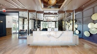 Located at 4310 Dunlavy St in Houston's Montrose neighborhood, this sophisticated two-floor space serves as a striking common area and lobby. The ground floor welcomes visitors through floor-to-ceiling glass entry doors, revealing polished travertine tile flooring that extends throughout. A sleek marble-fronted reception desk anchors the main lobby, set against a dramatic dark accent wall adorned with decorative circular art pieces. An ornate framed ceiling mural adds a refined touch overhead, while large windows invite abundant natural light and views of the surrounding greenery. A comfortable seating area with plush sofas and accent chairs provides an inviting lounge space. The lobby connects seamlessly to two hallways, one of which features a bold large-scale stained glass artwork reproduction, brass pendant lighting, and warm wood-toned tile flooring. A mirrored ceiling hallway leads to a stainless steel elevator and a grand staircase accented with brass railings and a striking sculptural chandelier. The upper floor expands the space considerably, housing additional hallways with wainscoting, panel molding, a beverage station with dark cabinetry, and a resident mailbox area. Throughout, the design balances classic architectural detailing with contemporary art and fixtures, creating a cohesive and upscale atmosphere.