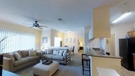 This single-floor residence is designed around an open-concept layout that connects the kitchen and living area, creating a spacious and cohesive atmosphere. Warm yellow-toned walls paired with light carpet flooring establish an inviting, airy ambiance throughout the main living space.

The living area is generously proportioned, accommodating a comfortable sectional-style sofa arrangement centered around a striped ottoman coffee table. A ceiling fan with a light fixture overhead provides both airflow and ambient lighting, while a track lighting system along the ceiling offers additional illumination. Crown molding adds a refined architectural detail along the upper walls.

Large windows dressed with vertical blinds allow ample natural light to fill the room. A glimpse into an adjoining room suggests additional living or sleeping quarters accessible directly from the main space.

The kitchen occupies the right side of the open floor plan, featuring dark countertops, white upper cabinetry, a built-in range hood, and a raised breakfast bar with seating. The seamless flow between the kitchen and living area makes this residence well-suited for both everyday living and entertaining.