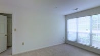 This single-floor space presents a clean, minimalist aesthetic defined by neutral tones and a calm, understated ambiance. The interior is characterized by smooth white walls that create a bright, open atmosphere, complemented by plush wall-to-wall carpeting in a warm beige tone that adds comfort underfoot. A centrally located window fitted with horizontal blinds allows natural light to filter into the room, illuminating the space and offering a glimpse of lush greenery outside. The ceiling is finished with a standard overhead vent for climate control, and a fire suppression sprinkler head is discreetly integrated into the ceiling surface, reflecting attention to safety standards. Electrical outlets and wall plates are positioned thoughtfully along the baseboards, maintaining the clean lines of the room. The crisp white baseboard trim provides a polished border between the walls and carpet, reinforcing the space's tidy, well-maintained appearance. Overall, the space offers a functional and versatile interior ready to accommodate a variety of uses, with its neutral palette and natural light serving as a welcoming foundation for personalization.