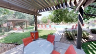 This expansive outdoor space showcases a harmonious blend of natural beauty and thoughtful design. The centerpiece is a sparkling swimming pool, surrounded by vibrant orange lounge chairs that add a pop of color to the serene environment. Multiple wooden pergolas provide shaded areas for comfortable seating, including a dining area with a metal table and matching orange chairs.

The landscaping is a testament to careful planning, with lush green lawns contrasting against areas of drought-resistant rock gardens. Mature trees, including pines and deciduous varieties, offer natural shade and privacy. Well-manicured shrubs and bushes line the perimeter, creating a sense of seclusion.

The space is designed for versatility, with ample room for both active recreation and peaceful relaxation. Concrete pathways wind through the area, connecting various zones and ensuring easy navigation. The overall ambiance is one of resort-like luxury, with the red brick buildings visible in the background adding a touch of classic architectural charm.

This outdoor oasis appears to be part of a larger complex, possibly an upscale apartment community or a high-end hotel, offering residents or guests a private retreat for sunbathing, swimming, and socializing in a meticulously maintained natural setting.