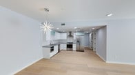 This single-floor space offers a clean, contemporary design highlighted by an open-plan kitchen and living area. The kitchen is outfitted with crisp white cabinetry, dark countertops, and a full suite of stainless steel appliances, including a gas range with a wall-mounted stainless hood, a built-in oven, and a French door refrigerator with an ice and water dispenser. Recessed lighting is complemented by a striking sputnik-style chandelier that serves as a focal point overhead, adding a bold, modern touch to the space. Light-toned hardwood flooring runs throughout, lending warmth and continuity to the interior. The walls are painted in a soft, neutral gray-white tone, contributing to the bright and airy atmosphere. A hallway visible toward the rear of the space suggests additional rooms beyond the main open area. The exterior features a private patio enclosed by tan-toned cinder block walls, with established greenery and tropical plantings along the perimeter, providing a sense of seclusion while allowing open sky views above. The coated concrete patio floor adds a finished, functional quality to the outdoor space.