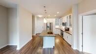 This single-floor residence offers a thoughtfully designed open layout that balances comfort and functionality throughout. The kitchen serves as a central highlight, featuring a large center island with a dark stone countertop and undermount sink, crisp white shaker-style cabinetry with glass-front upper panels, stainless steel appliances including a French door refrigerator, electric range, and built-in microwave, all illuminated by elegant pendant lighting. Rich hardwood-style flooring extends seamlessly through the kitchen and living areas. The living room is bright and welcoming, centered around a sliding glass door that opens to a private balcony with outdoor views, complemented by a ceiling fan overhead. Two well-sized bedrooms feature plush carpet flooring, ceiling fans, and windows that bring in natural light. The primary bedroom connects to a private en-suite bathroom with dual undermount sinks, dark stone countertops, wood-look flooring, and direct access to a spacious walk-in closet fitted with white wire shelving. A second full bathroom offers a tiled tub-shower surround, a white vanity with stone counters, and wood-look flooring. The walk-in closet provides ample organization space with multi-level wire shelving. Neutral warm tones and clean finishes throughout create a cohesive, modern aesthetic.