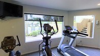 This single-floor fitness room is designed to provide a functional and comfortable workout environment. The space is outfitted with modern cardio equipment, including an elliptical trainer and a treadmill, positioned to take full advantage of the room's large windows. These expansive windows allow abundant natural light to fill the space while offering clear views of the surrounding outdoor area, which features a pool, fencing, and lush greenery — creating an pleasant backdrop for exercise. The walls are finished in a clean, neutral tone, contributing to a bright and open atmosphere. A roller shade on the primary window provides flexibility in light control when needed. The layout is practical and uncluttered, allowing for ease of movement between equipment. A wall-mounted screen is also visible, adding a functional entertainment element to the workout experience. The overall design of the space prioritizes utility and comfort, making it a well-suited facility for residents or guests looking for a convenient place to maintain an active lifestyle.