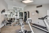 Located at 27550 Hillcrest in Mission Viejo, California, this single-floor fitness center is a well-equipped community exercise facility designed to support a well-rounded workout routine. The space features vaulted ceilings that contribute to an open, airy feel, complemented by a combination of overhead lighting and natural light filtering through glass block windows and large sliding glass doors.

The cardio section is outfitted with a treadmill, elliptical trainers, and stationary bikes, all positioned to take advantage of the wall-mounted television and views of the outdoor pool area beyond the sliding glass doors. A seating area near the entrance provides a convenient spot to rest between sessions.

The strength training area is lined with floor-to-ceiling mirrors that visually expand the space and allow for proper form monitoring. Equipment includes a multi-function cable machine, a weight bench, barbells, a curl bar, and a selection of dumbbells and free weights. Wall-mounted fans supplement the HVAC system to maintain a comfortable workout environment.

Carpeted flooring with protective exercise mats underfoot runs throughout the space, and posted rules and regulations ensure an organized, community-friendly atmosphere. The direct sightline to the pool and patio area, complete with umbrella-shaded tables, adds an inviting outdoor connection to this functional fitness facility.