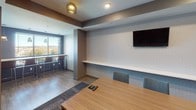 This contemporary meeting space is thoughtfully designed to support professional gatherings and collaborative work. The room's most striking architectural feature is a floor-to-ceiling slatted wood accent wall, crafted from rich, warm-toned timber planks arranged horizontally to create a bold visual statement and natural texture against the cool grey and blue-grey wall finishes throughout.

The main meeting area centers around a long, light wood conference table surrounded by upholstered blue-grey chairs, comfortably accommodating a sizeable group. Built-in connectivity features are integrated directly into the table surface. Opposite the accent wall, a wall-mounted flat-screen television is positioned above a continuous floating white shelf, providing a functional media and presentation focal point ideal for meetings and presentations.

Adjacent to the main conference area, a windowed bar-height counter offers an additional gathering spot with tall stools, allowing natural light to filter into the space through slatted window blinds. The flooring transitions between warm wood-look planks and a patterned carpet tile in the seating zone, adding definition to the layout.

Recessed and flush-mount ceiling fixtures provide balanced ambient lighting throughout, complemented by subtle pendant accents. A secondary open area beyond the main room offers flexible overflow or breakout space. The overall ambiance is polished, functional, and modern.