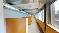 This single-floor commercial office space blends industrial and contemporary design elements to create a functional and visually distinctive work environment. The main open-plan area is defined by a row of built-in maple wood workstations with dark countertops, arranged in an elongated corridor-style layout alongside a continuous bank of floor-to-ceiling windows that flood the space with natural light and offer views of the surrounding exterior. Polished terrazzo floors run throughout the open area, adding a refined touch to the industrial aesthetic. Overhead, exposed concrete ceilings showcase prominent silver HVAC ductwork and structural beams, along with visible conduit and utilities—a nod to the building's raw architectural character. At the far end of the open floor, a dedicated conference room provides a more private setting, furnished with an oval table and ergonomic white chairs. The room features warm yellow accent walls, built-in maple cabinetry, and a wall-mounted flat-screen display, making it well-suited for meetings and presentations. Glass partitions and concrete structural columns further define the spatial transitions throughout the office. The overall ambiance is professional yet creatively inspired, combining warm wood tones, bold accent colors, and exposed industrial materials for a dynamic workplace setting.