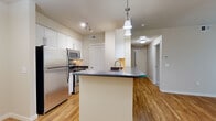 Located at 451 Westcliffe Blvd in Richland, WA, this one-bedroom, one-bathroom unit offers a well-designed single-floor layout that makes efficient use of its space. The open-concept kitchen, dining, and living area serves as the heart of the unit, anchored by a central kitchen island with dark countertops and pendant lighting overhead. The kitchen is equipped with stainless steel appliances — including a refrigerator, range, and microwave — along with white cabinetry, a double-basin sink, and a built-in dishwasher. Warm hardwood-style flooring runs throughout the main living areas, lending a cohesive and inviting feel. The living and dining areas provide an open, flexible space with neutral tones and clean lines. A hallway connects the main area to the private bedroom, which features plush carpeting, vertical blinds, and windows that allow natural light to filter in. The bathroom includes a vanity with dark countertop, an undermount sink, a large mirror, and under-sink storage. The unit's neutral color palette and consistent finishes throughout create a clean and functional living environment suitable for a range of lifestyles.