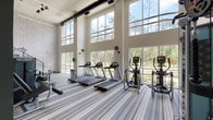 This two-story fitness center is designed to inspire an energizing workout experience through its open, airy architecture and abundance of natural light. The main floor is anchored by a dramatic wall of floor-to-ceiling windows that frame sweeping views of surrounding greenery and manicured grounds, flooding the space with daylight and creating a bright, motivating atmosphere. Striped commercial carpeting runs throughout, complementing the modern, neutral color palette of white walls and dark structural accents.

The main level is generously equipped with a row of treadmills and elliptical machines positioned directly in front of the expansive windows, allowing users to enjoy outdoor views during cardio workouts. Matrix-brand strength-training machines, including a functional trainer and cable systems, are centrally positioned alongside adjustable benches and a dedicated free weights area with a mirrored wall. A water cooler station adds convenience to the workout floor.

A glass-railed staircase connects the main floor to the upper mezzanine level, which overlooks the primary workout area below through sleek railings, offering additional workout space. Exposed industrial-style ceiling beams, track lighting, and wall-mounted fans complete the contemporary design, blending functionality with a polished aesthetic throughout.