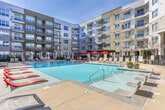 This expansive outdoor amenity space is situated within a modern mid-rise community in Atlanta's 30324 corridor. At its centerpiece is a generously sized swimming pool with crystal-clear blue water, framed by a broad concrete pool deck outfitted with rows of cushioned lounge chairs in warm terracotta tones, ideal for sunbathing or relaxing. Decorative potted plantings with seasonal flowers accent the poolside, adding color and a resort-like atmosphere.

Adjacent to the pool, a dedicated cabana zone features multiple white-draped canopy structures set on a raised artificial turf platform, providing shaded, semi-private retreats. The cabanas are complemented by bright orange accent furniture, contributing a vibrant, contemporary aesthetic.

On the opposite side of the courtyard, a well-appointed outdoor entertaining area includes a built-in grilling station, a covered pergola strung with bistro lighting, and a mix of bar-height and standard dining tables with red umbrellas. The multi-story building façade, featuring large windows and private balconies, encloses the courtyard on multiple sides, creating an intimate, sheltered atmosphere throughout this amenity-rich outdoor space.