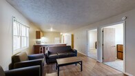 This single-floor unit is designed with a practical and functional layout. The living room serves as the central gathering space, featuring rich hardwood-style flooring, a textured ceiling, and baseboard heating. A front entry door with small window panels allows natural light into the room, which comfortably accommodates a seating arrangement centered around a wood coffee table.

The kitchen is generously sized and open in feel, offering warm-toned wooden cabinetry, light granite-style countertops, and a full suite of white appliances including a refrigerator, electric range, dishwasher, and microwave. A kitchen bar with seating connects this space to the living area, making it ideal for casual dining or entertaining.

The unit includes multiple bedrooms, each carpeted and furnished with a bed frame, dresser, and desk, providing a functional setup for residents. Windows in each room allow natural light to filter in. One bedroom features hardwood-style flooring and a baseboard heater.

Two bathrooms serve the unit, each equipped with an oak-toned vanity, mirror, and light fixture. One bathroom includes a shower/tub combination, while the other provides a more compact layout. Both feature hardwood-style flooring and clean, neutral finishes throughout.