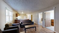 This single-floor unit is designed with a practical and functional layout. The living room serves as the central gathering space, featuring rich hardwood-style flooring, a textured ceiling, and baseboard heating. A front entry door with small window panels allows natural light into the room, which comfortably accommodates a seating arrangement centered around a wood coffee table.

The kitchen is generously sized and open in feel, offering warm-toned wooden cabinetry, light granite-style countertops, and a full suite of white appliances including a refrigerator, electric range, dishwasher, and microwave. A kitchen bar with seating connects this space to the living area, making it ideal for casual dining or entertaining.

The unit includes multiple bedrooms, each carpeted and furnished with a bed frame, dresser, and desk, providing a functional setup for residents. Windows in each room allow natural light to filter in. One bedroom features hardwood-style flooring and a baseboard heater.

Two bathrooms serve the unit, each equipped with an oak-toned vanity, mirror, and light fixture. One bathroom includes a shower/tub combination, while the other provides a more compact layout. Both feature hardwood-style flooring and clean, neutral finishes throughout.
