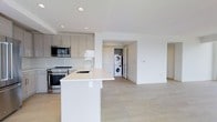 This contemporary single-floor space showcases a clean, modern aesthetic defined by an open-concept layout and a bright, neutral palette throughout. The kitchen is a standout feature, equipped with stainless steel appliances including a range, microwave, and a deep single-basin sink set into a crisp white countertop. Soft-toned cabinetry with a matte finish lines the walls, providing generous storage while maintaining a streamlined look. A central kitchen island extends the counter space and naturally delineates the kitchen from the adjoining living area, creating a cohesive and functional flow. Recessed lighting in the kitchen area illuminates the space with even, ambient light. The flooring throughout is a light-toned wood-look material that adds warmth while complementing the overall minimalist design. A dedicated in-unit laundry area is conveniently tucked away, housing a stacked washer and dryer unit within its own closeted space. The open living area beyond the kitchen offers a blank canvas with clean white walls and smooth ceilings, providing a flexible and airy environment. The overall ambiance is fresh, modern, and move-in ready, reflecting a thoughtfully designed and highly functional living space.