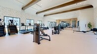 Located at 25440 Solaris Pl in Murrieta, CA, this single-floor fitness center offers a generously sized, thoughtfully designed exercise facility that caters to a wide range of workout preferences. The space is defined by soaring ceilings accented with rustic wood beams, crisp white walls, and a combination of light wood-tone and neutral flooring that creates a clean, welcoming atmosphere throughout.

Large black-framed windows and glass doors line the perimeter, flooding the interior with natural light and offering views of the surrounding outdoor landscape, including palm trees and a pool area. A striking textured stone accent wall serves as a standout architectural feature, adding warmth and visual interest.

The facility is comprehensively equipped with cardio machines including treadmills, ellipticals, stationary bikes, and rowing ergometers. A dedicated strength training zone houses cable machines, multi-functional racks, free weights, and adjustable benches, complemented by full-length mirrors and warm wood cabinetry. A highlight of the space is an indoor climbing wall, providing a unique functional fitness option. High ceilings throughout accommodate tall cable and functional training rigs with ease. The overall ambiance is modern yet inviting, blending upscale design with practical, high-performance fitness infrastructure.