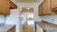 This single-floor residence offers a practical and comfortable layout designed for everyday living. The kitchen is well-appointed with warm honey-toned wood cabinetry, granite-look countertops, and white appliances including a full-size refrigerator and dishwasher. Ample cabinet storage lines both sides of the kitchen, providing generous space for organization and meal preparation. An open archway connects the kitchen directly to the living area, creating a seamless flow between the two spaces and enhancing the sense of openness throughout the home. The living area is bathed in natural light streaming through a large window, illuminating the beautifully maintained hardwood floors that run throughout. The warm wood tones of the flooring lend a classic, inviting character to the space. A wall-mounted climate control unit ensures year-round comfort. The neutral wall tones throughout provide a versatile backdrop, making the space easy to personalize. The overall ambiance is clean, bright, and functional, offering a welcoming environment that balances practicality with a timeless residential aesthetic.