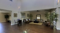 This single-floor space showcases a thoughtfully designed open-concept layout that seamlessly connects its living and dining areas into one expansive, flowing environment. The most striking architectural feature is the dramatic vaulted ceiling, which rises impressively over the main living zone, creating a grand sense of volume and openness. Warm-toned hardwood flooring runs throughout, grounding the space with a rich, cohesive aesthetic.

The dining area is positioned toward the front, furnished with a glass-top table and sleek dark chairs, offering an intimate yet stylish setting. A sculptural wall sconce provides soft accent lighting, complementing the neutral taupe and beige wall tones that lend the space a calm, sophisticated ambiance.

The living area is generously proportioned and features two distinct seating arrangements — a plush, dark sectional sofa and a lighter-toned sofa — both anchored by a natural fiber area rug. A central coffee table with a decorative tabletop fountain adds a refined focal point. Tall potted bamboo plants are positioned throughout, introducing organic texture and a sense of tranquility. A ceiling fan with integrated lighting serves the deeper lounge area. A large decorative mirror reflects light and enhances the sense of depth, while a partial view into an adjacent room suggests additional functional spaces beyond.