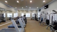 This fitness center offers a well-organized, single-floor layout designed to accommodate a variety of workout routines. The space is outfitted with multiple treadmills and elliptical machines arranged along one side, providing ample cardio options for users. Strength training is supported by a cable/pulley multi-station machine, a dedicated weight bench with a barbell rack, and additional resistance equipment positioned along the perimeter.

The room features neutral-toned walls paired with a beige carpeted floor, contributing to a clean and functional aesthetic. Recessed lighting is distributed evenly across the ceiling, ensuring the entire space is well-lit during any hour of use. A ceiling fan provides additional air circulation, enhancing comfort during workouts. A wall-mounted television is positioned on one side of the room, adding an entertainment element for users.

Several windows fitted with blinds line the walls, allowing natural light to filter into the space while maintaining privacy. A large mirror is mounted along one wall, a practical addition that aids in monitoring exercise form. The overall layout is open and uncluttered, allowing for easy movement between equipment stations.