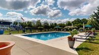 Located at 5398 Rockingham Ct in Westerville, Ohio, this community recreational area centers around a generously sized outdoor swimming pool that serves as the focal point of the space. The pool is surrounded by a wide concrete sundeck lined with lounge chairs, offering ample room for relaxation and sunbathing under open skies.

The pool area is well-appointed with multiple shaded seating zones, including umbrella-covered tables and a pergola-style canopy, providing comfortable spots for gathering outside of the water. Lush green lawns border the deck, complemented by mature trees — including a graceful weeping willow — that add natural character and shade to the environment.

A dedicated pool house structure, finished in white painted brick with a pitched roof, provides essential amenities including safety equipment mounted on the exterior wall and pool rules signage. A children's playground is visible beyond the fencing, making this a family-friendly recreational destination.

The entire area is enclosed by black iron fencing, ensuring a secure environment. Decorative potted plants and flower arrangements are placed throughout, adding color and charm. The space benefits from abundant natural light and open sky views, creating an inviting outdoor atmosphere ideal for warm-weather community recreation.