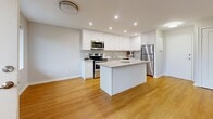 This 2-bedroom, 1-bathroom single-floor residence offers a clean, modern aesthetic with a practical and efficient layout. Warm hardwood-style flooring runs consistently throughout every room, creating a cohesive and inviting atmosphere.

The kitchen is a standout feature, outfitted with crisp white shaker cabinetry extending to the ceiling, stone-look countertops, a subway tile backsplash, and a full suite of stainless steel appliances including a range, built-in microwave, and refrigerator. A central island provides additional prep space and opens to the adjacent living area, which benefits from large windows that invite natural light and offer views of the surrounding neighborhood.

Both bedrooms are well-proportioned and feature double-door closets, neutral wall tones, and windows that bring in natural light. The primary bedroom is notably spacious, offering generous built-in closet storage. The second bedroom also includes a closet and a bright window overlooking a green outdoor view.

The bathroom is finished with a granite-topped white vanity, an undermount sink, and a chrome fixture, reflecting the same clean and contemporary style found throughout the unit. Additional storage is provided by a dedicated closet. The overall ambiance is bright, neutral, and move-in ready.