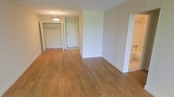 This single-floor space offers a clean, functional layout designed to maximize usability. The main living area is finished with warm-toned hardwood-style flooring that extends throughout, providing a cohesive and inviting atmosphere. Neutral, light-toned walls create a bright and versatile backdrop, allowing the space to feel open and airy.

A pendant light fixture is centered near the entryway, offering both practical illumination and a subtle decorative touch. Just inside the front door, an open closet with a visible hanging rod provides convenient storage for coats, shoes, or additional belongings. The entryway transitions naturally into the main living area, which offers an open feel well-suited for flexible furniture arrangements.

A partial wall defines one side of the living space, adding a sense of structure without closing off the area. A bathroom is accessible directly from this zone, as indicated by visible fixtures. The combination of light flooring, white trim, and pale walls gives the overall space a clean, move-in-ready aesthetic that is both practical and welcoming.