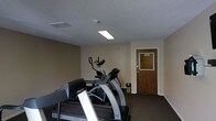 This single-floor fitness room provides a dedicated space for cardiovascular exercise and general wellness. The room is outfitted with multiple cardio machines, including Precor-brand treadmills and an elliptical trainer, making it well-suited for a variety of workout routines. The space features neutral beige walls, dark carpeted flooring, and a textured ceiling with flush-mounted lighting that provides adequate illumination throughout. Ventilation is supported by a ceiling air vent, ensuring a comfortable environment during physical activity. A solid wood-panel door with a windowed upper panel serves as the main entry point, and a small waste bin is conveniently placed near the equipment. The room's layout is straightforward and functional, maximizing the use of available floor space for exercise equipment. While modest in scale, the fitness room is thoughtfully arranged to accommodate users comfortably. Overall, the space delivers a clean, practical environment focused entirely on providing accessible fitness amenities.