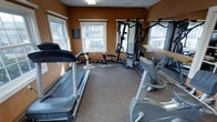 This single-floor fitness room is designed to support a well-rounded workout routine in a compact yet functional space. The room features warm terracotta-toned walls that create an energizing atmosphere, complemented by natural light streaming in through multiple double-hung windows that offer views of the surrounding area.

The flooring consists of durable, cushioned exercise-grade carpeting, providing comfort and stability underfoot during workouts. The space is thoughtfully arranged to maximize usability, housing a treadmill and an upright stationary bike for cardiovascular training. At the center of the room stands a multi-station cable and weight machine, offering a wide variety of strength training exercises. A set of free weights and an adjustable bench are also available for supplemental resistance training.

A pull-up and dip bar station is mounted along one wall, further expanding the range of bodyweight exercises available. The room is well-lit with overhead fluorescent lighting in addition to the natural light provided by the windows. A posted notice board is visible on one wall, likely displaying fitness guidelines or facility rules. Overall, the space is clean, organized, and outfitted to meet the fundamental fitness needs of its users.