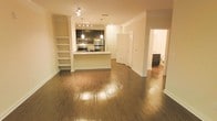 This two-floor space showcases a well-designed open-concept main level that seamlessly connects the living area and kitchen. Rich hardwood flooring flows throughout, reflecting warm ambient lighting and adding a polished, cohesive feel to the space.

The kitchen is a functional centerpiece, featuring dark cabinetry, stainless steel appliances including a refrigerator, microwave, and range, as well as a peninsula with a light-toned countertop that provides additional prep space and casual seating potential. Pendant lighting hangs above the peninsula, complemented by a track lighting fixture that illuminates the kitchen area effectively.

A set of built-in open shelves is positioned along one wall adjacent to the living space, offering practical display or storage options. Crown molding lines the ceilings, adding a refined architectural detail throughout the main level. A hallway visible from the main area leads to additional rooms, including what appears to be a bathroom, suggesting a thoughtful layout that balances open living with private spaces. The overall ambiance is warm and inviting, with clean lines and neutral tones creating a versatile backdrop.